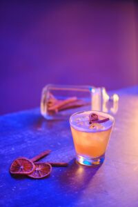 Close-up of a glass cocktail with cinnamon and dried orange slices on a colorful surface.